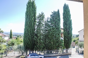 Área residencial com ciprestes altos, um carro estacionado e um edifício branco com telhado em cerâmica.