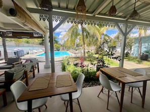 An outdoor patio with wooden tables and white chairs, overlooking a swimming pool and lush greenery.