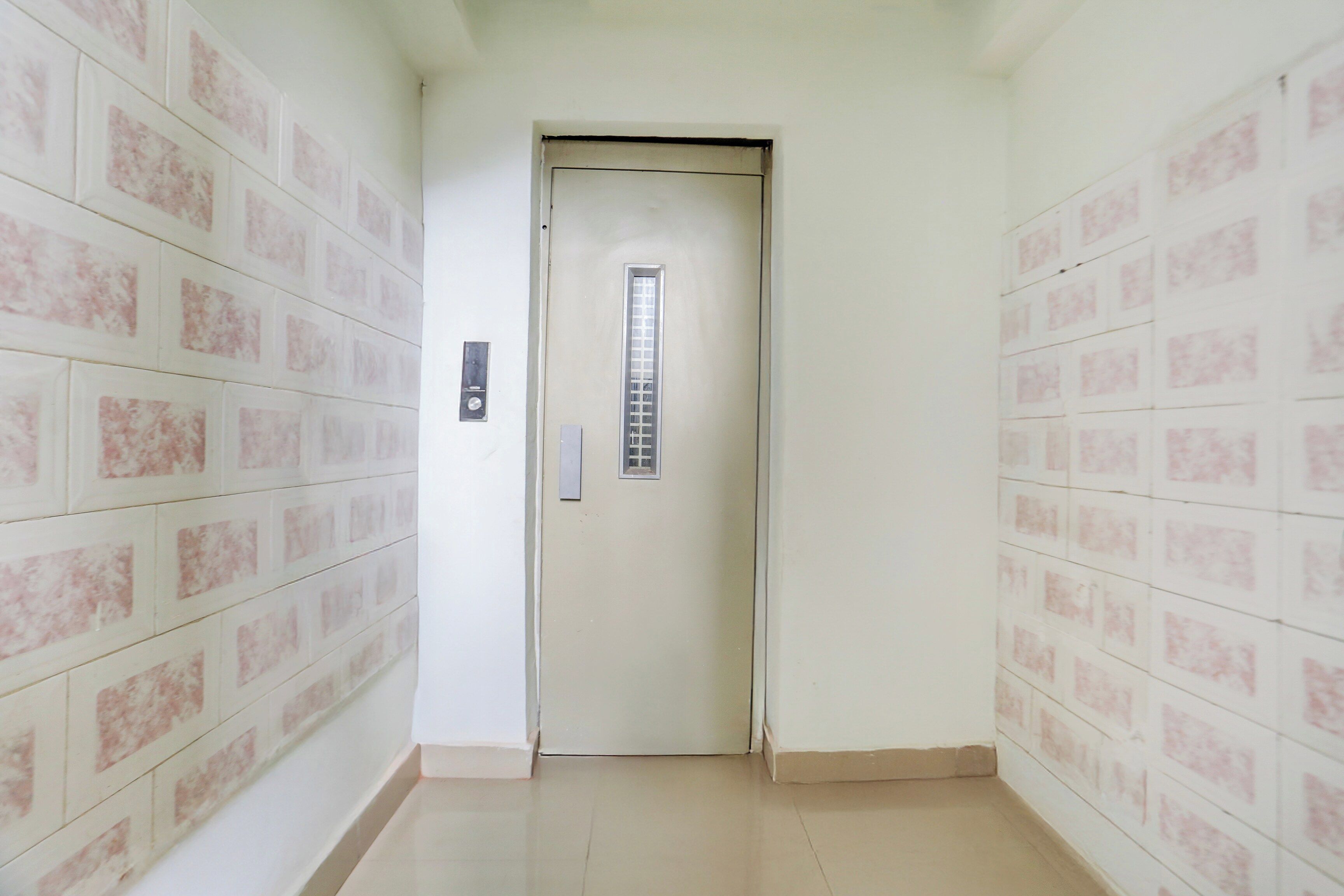Un pasillo estrecho con una puerta al final, un portero automático en la pared y un piso de baldosas.