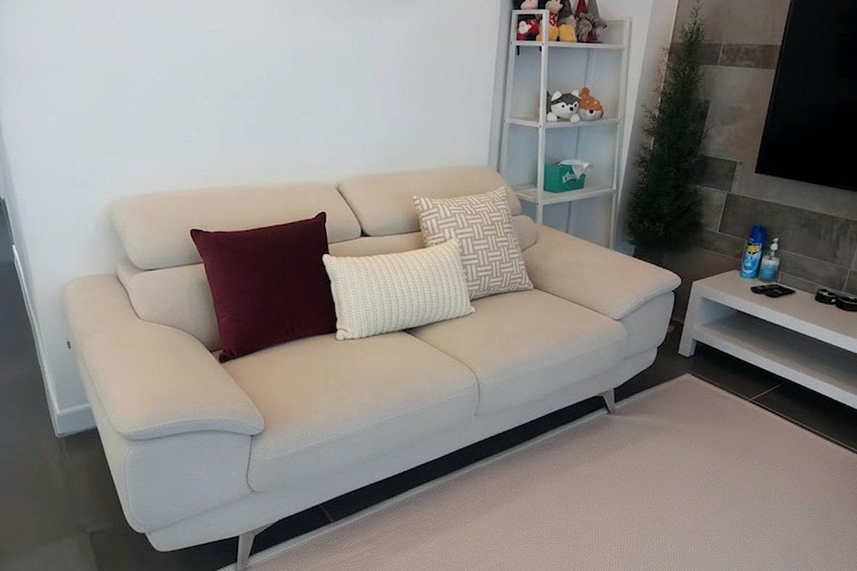 Un sofá color beige con dos almohadas, un mueble blanco para entretenimiento con juguetes, y una pequeña planta.