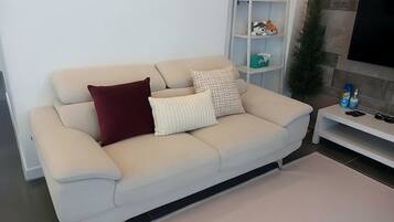 Un sofá color beige con dos almohadas, un mueble blanco para entretenimiento con juguetes, y una pequeña planta.
