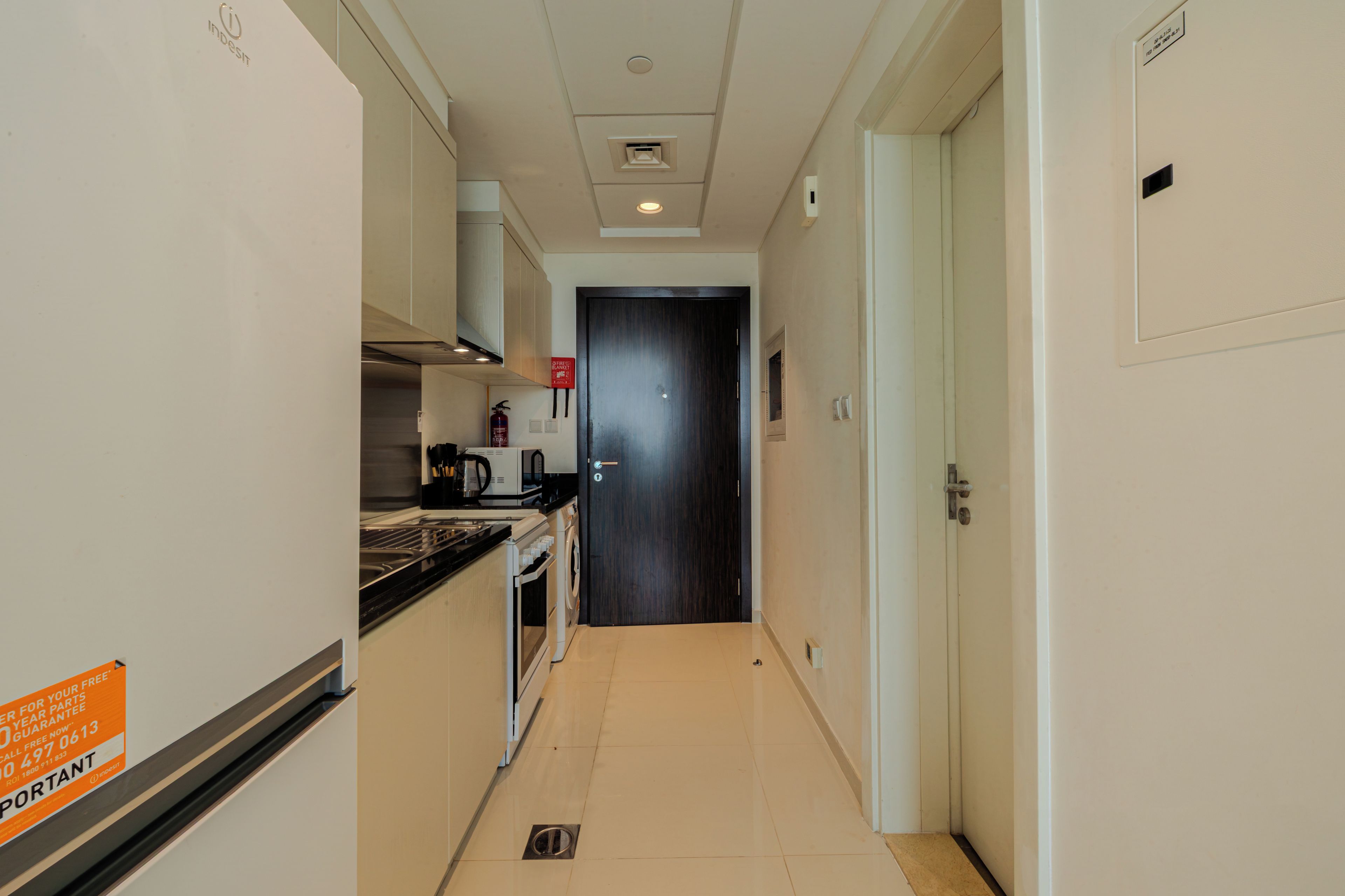 A narrow hallway with a kitchen area on one side, a dark door at the end, and a fire safety sign.