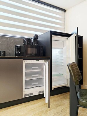 A compact kitchenette with a built-in refrigerator and microwave, a chair, and a window with blinds. - Ocean Paradise apartment (Mellieħa)