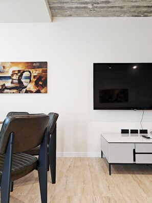 A modern living room with a flat-screen TV mounted on the wall, a dining area with a table and chairs, and a painting above the TV. - Ocean Paradise apartment (Mellieħa)