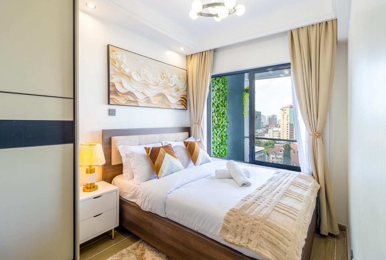 A modern hotel room with a large bed, a nightstand with a lamp, a city view through a window, and a decorative wall piece.