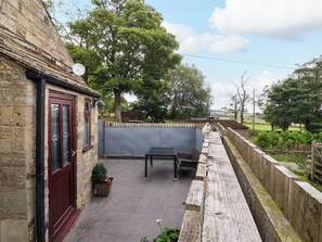 A stone building with a red door, a patio with a table and chairs, and a view of the countryside. - Brownhill Bungalow (Keighley)