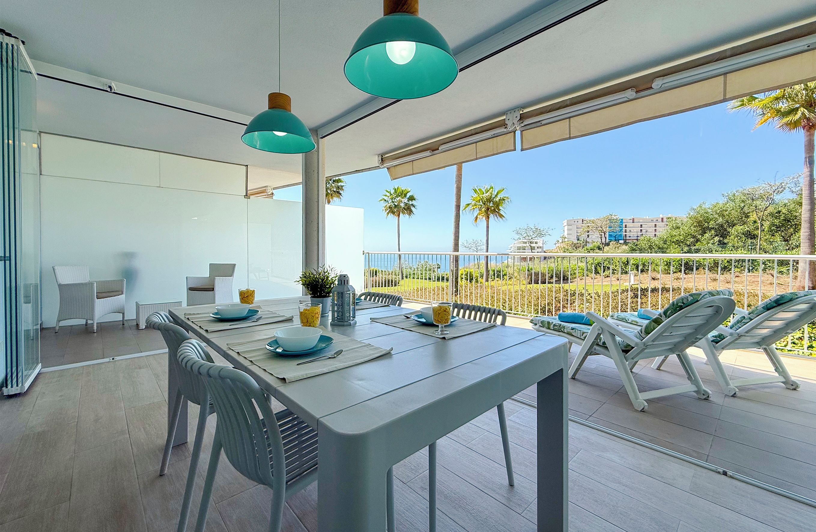 Un área de comedor moderna al aire libre con una mesa preparada para dos personas, rodeada de sillas y una vista de palmeras y edificios al fondo.