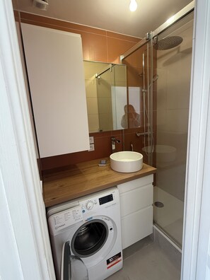 Une salle de bain compacte dotée d’une douche à parois de verre, d’un lave-linge encastré et d’un plan de travail en bois.