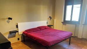 Ein Einzelbett mit Kopfteil, ein Nachttisch, ein Heizkörper und ein Fenster mit Jalousien.