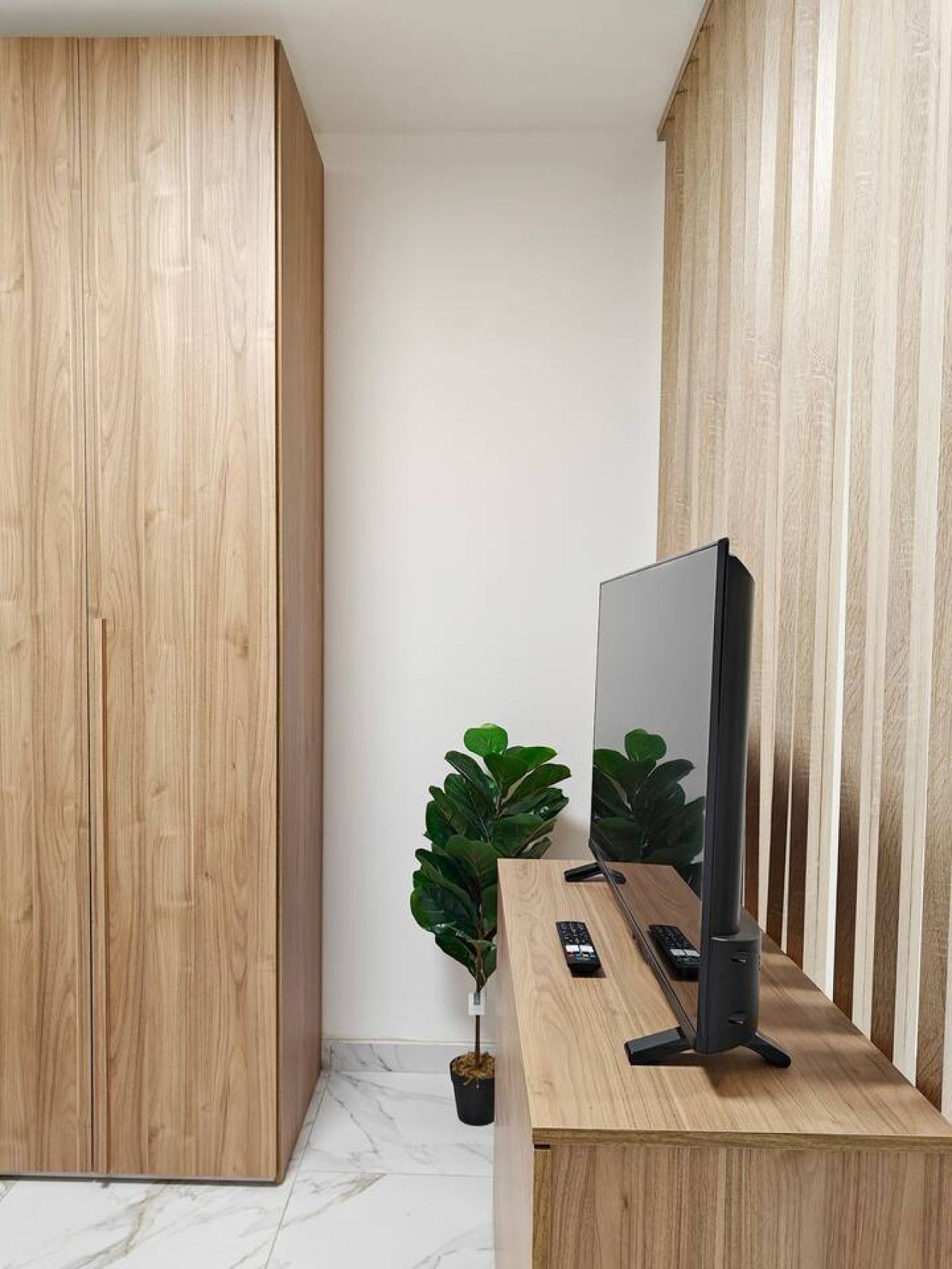 Une chambre moderne avec une armoire en bois, un bureau doté d’un téléviseur à écran plat, une télécommande et une plante en pot.