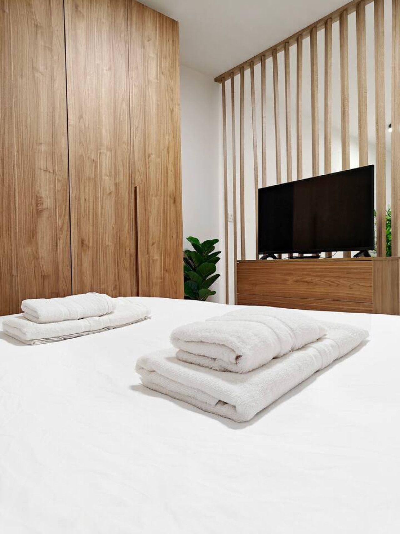 Un dormitorio moderno con un armario de madera, un televisor de pantalla plana grande y toallas blancas ordenadas sobre la cama.