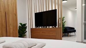 Habitación de hotel moderna con cama, televisor de pantalla plana montado en un panel de madera, planta en maceta y un sillón oscuro.