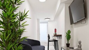 Una sala moderna con un sofá negro, una planta verde grande, piso de mármol, televisor de pantalla plana montado en la pared y un área de comedor al fondo.
