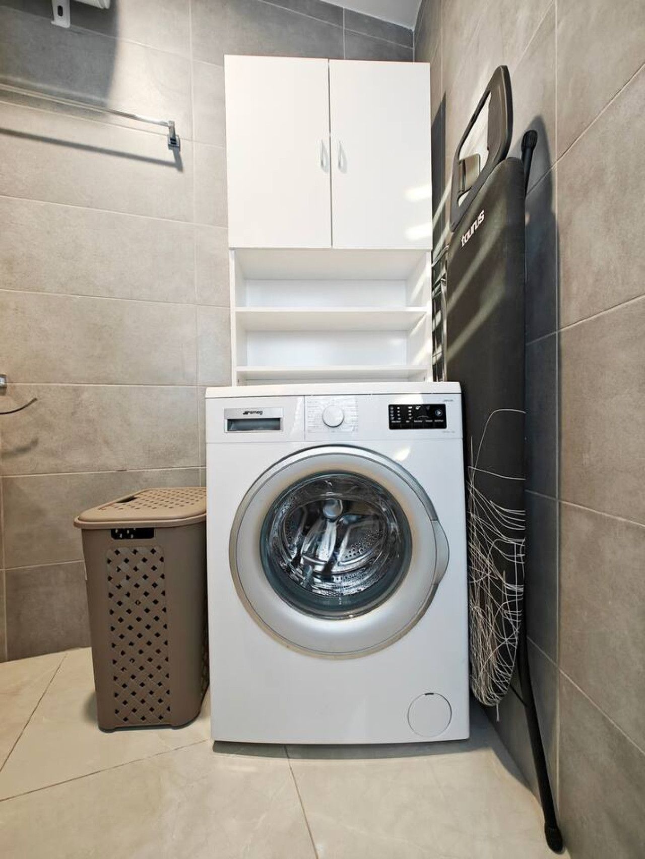A compact laundry area with a front-loading washing machine, a drying rack, and a storage cabinet.