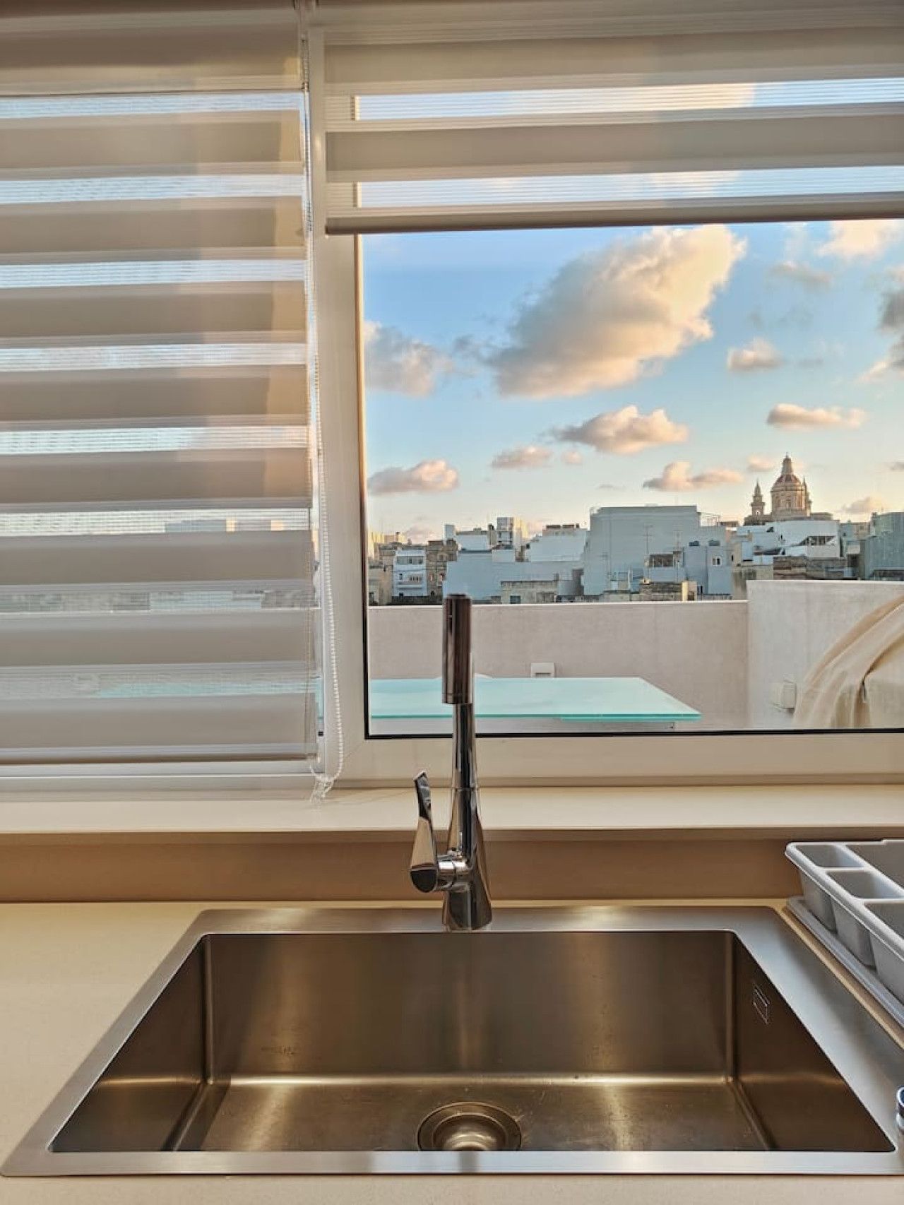 A modern kitchen with a stainless steel sink, a window with blinds, and a cityscape view.