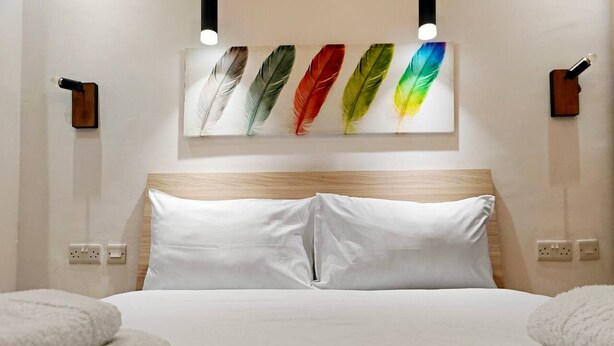 Quarto de hotel com cama, almofadas brancas, cabeceira, obra de arte colorida na parede com penas e duas luminárias fixadas na parede.