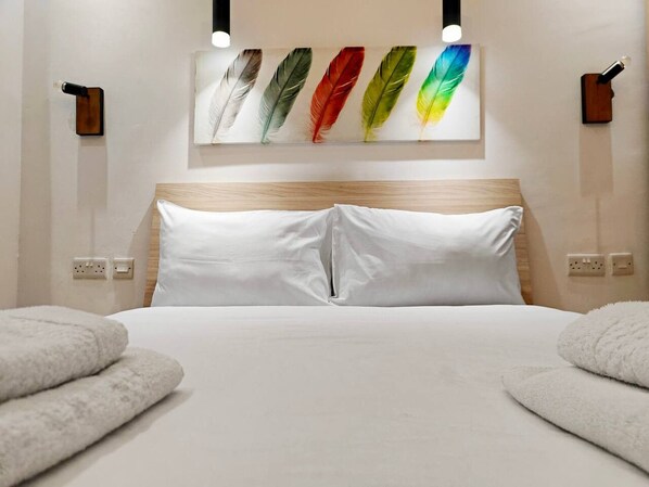 A hotel room with a bed, white pillows, a headboard, a wall-mounted artwork of colorful feathers, and two wall-mounted light fixtures.