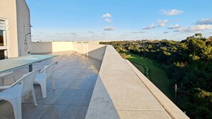 Terraço no telhado com mesa de vidro e cadeiras brancas, com vista para uma paisagem verdejante.
