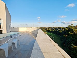 A rooftop terrace with a glass table and white chairs, overlooking a green landscape.