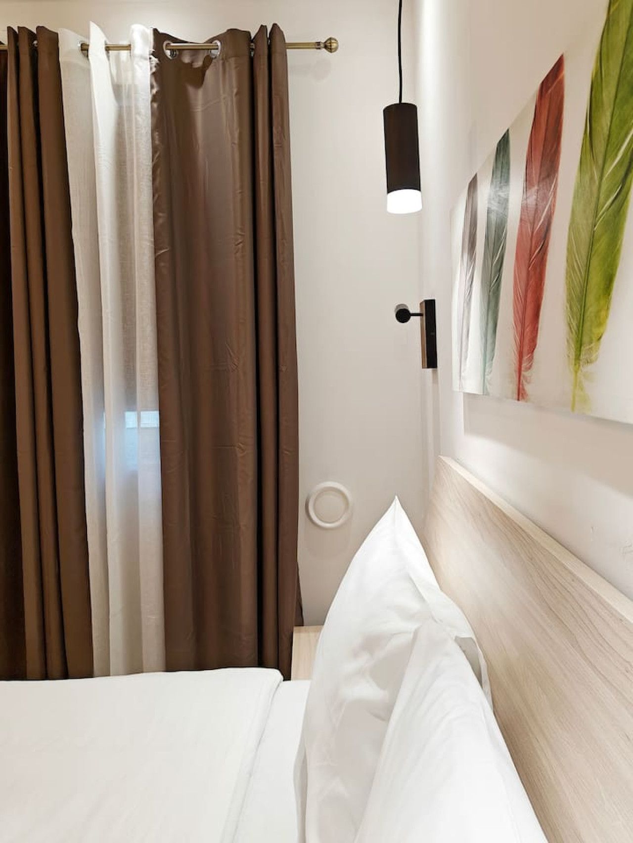 Habitación de hotel con cama, cortinas, una lámpara fijada a la pared y una obra de arte con plumas.