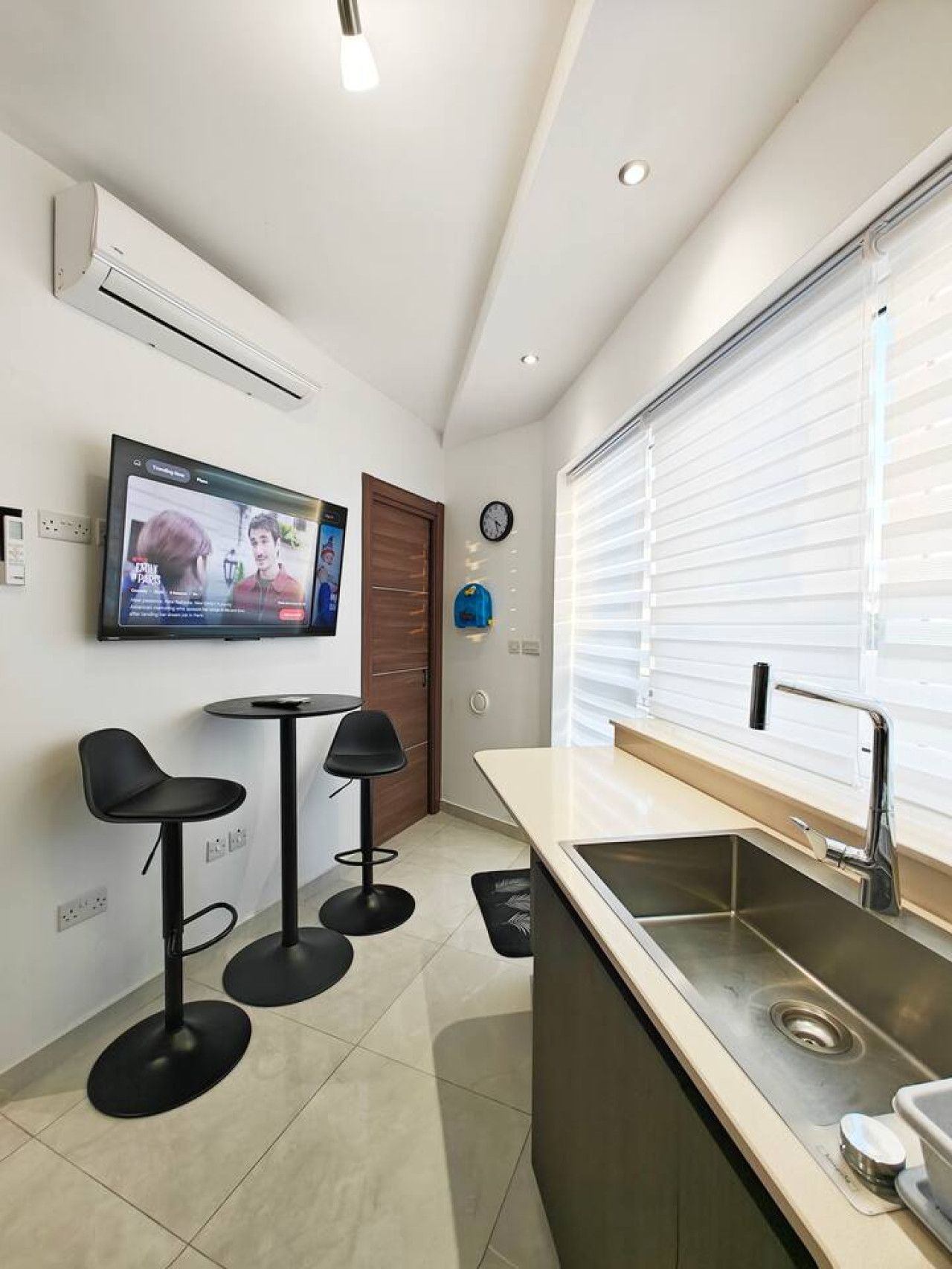 A modern kitchen with a built-in sink, a flat-screen TV mounted on the wall, and two black bar stools.
