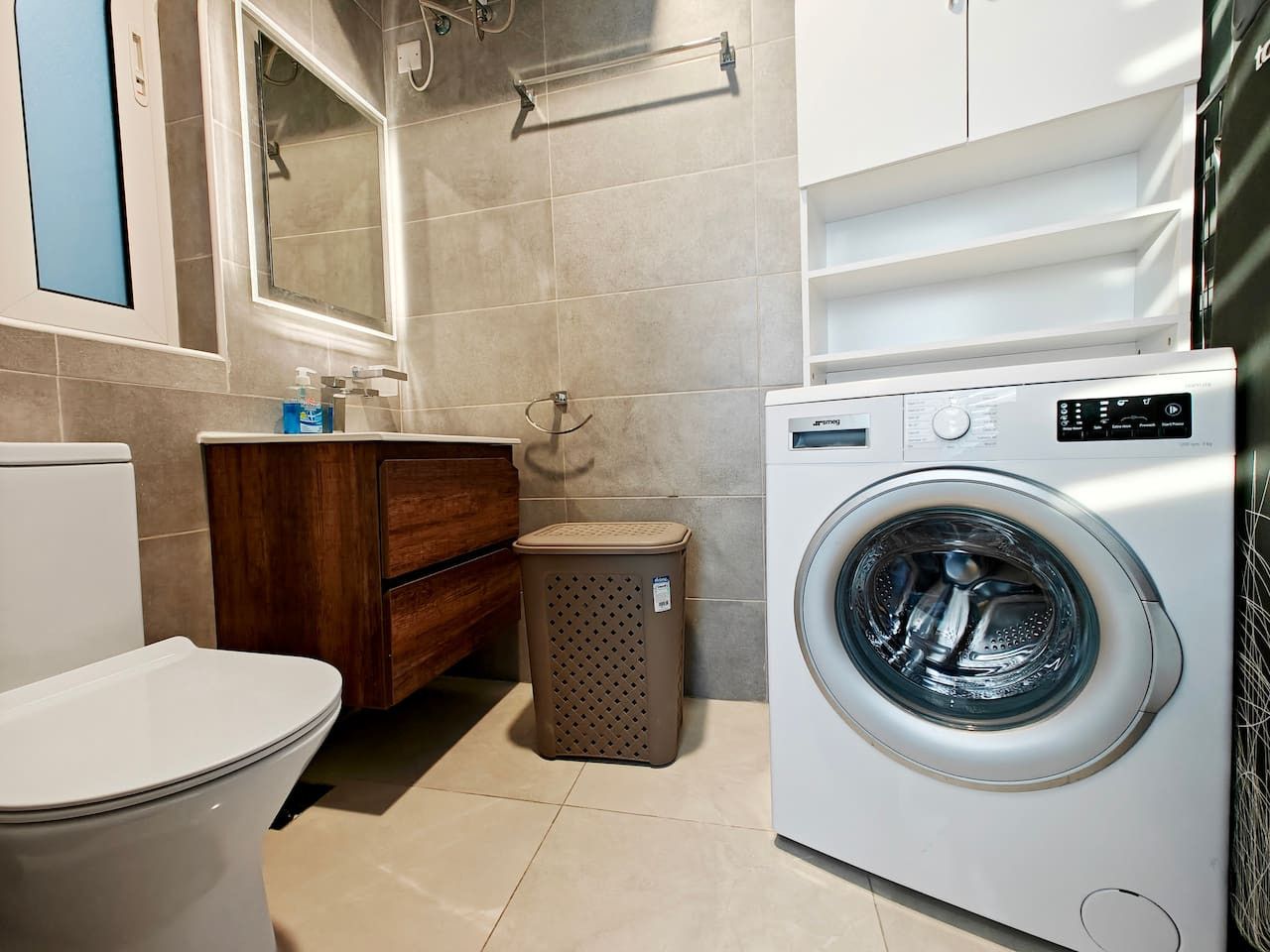 A bathroom with a toilet, a washing machine, a wooden cabinet, and a trash bin.