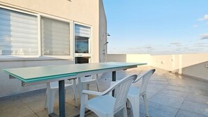 Um pátio externo com mesa e cadeiras, uma janela com persianas e um céu limpo.