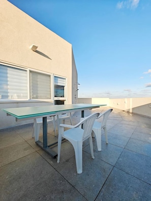 An outdoor patio with a table and chairs, a window with blinds, and a clear sky.