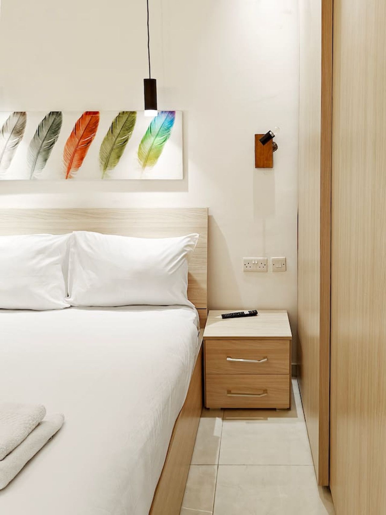 A hotel room with a bed, a bedside table with a remote control, a wall-mounted power outlet, and a door.