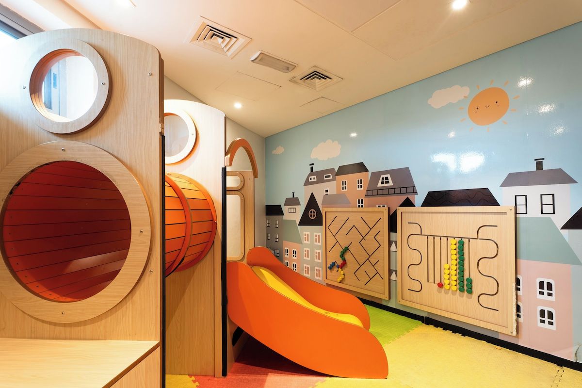 Un espace de jeux pour enfants avec des structures en bois de forme circulaire, un toboggan et une fresque murale représentant des maisons.
