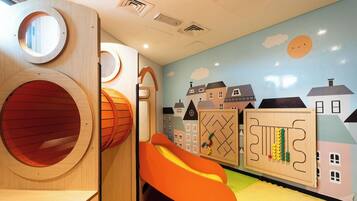 Un espace de jeux pour enfants avec des structures en bois de forme circulaire, un toboggan et une fresque murale représentant des maisons.