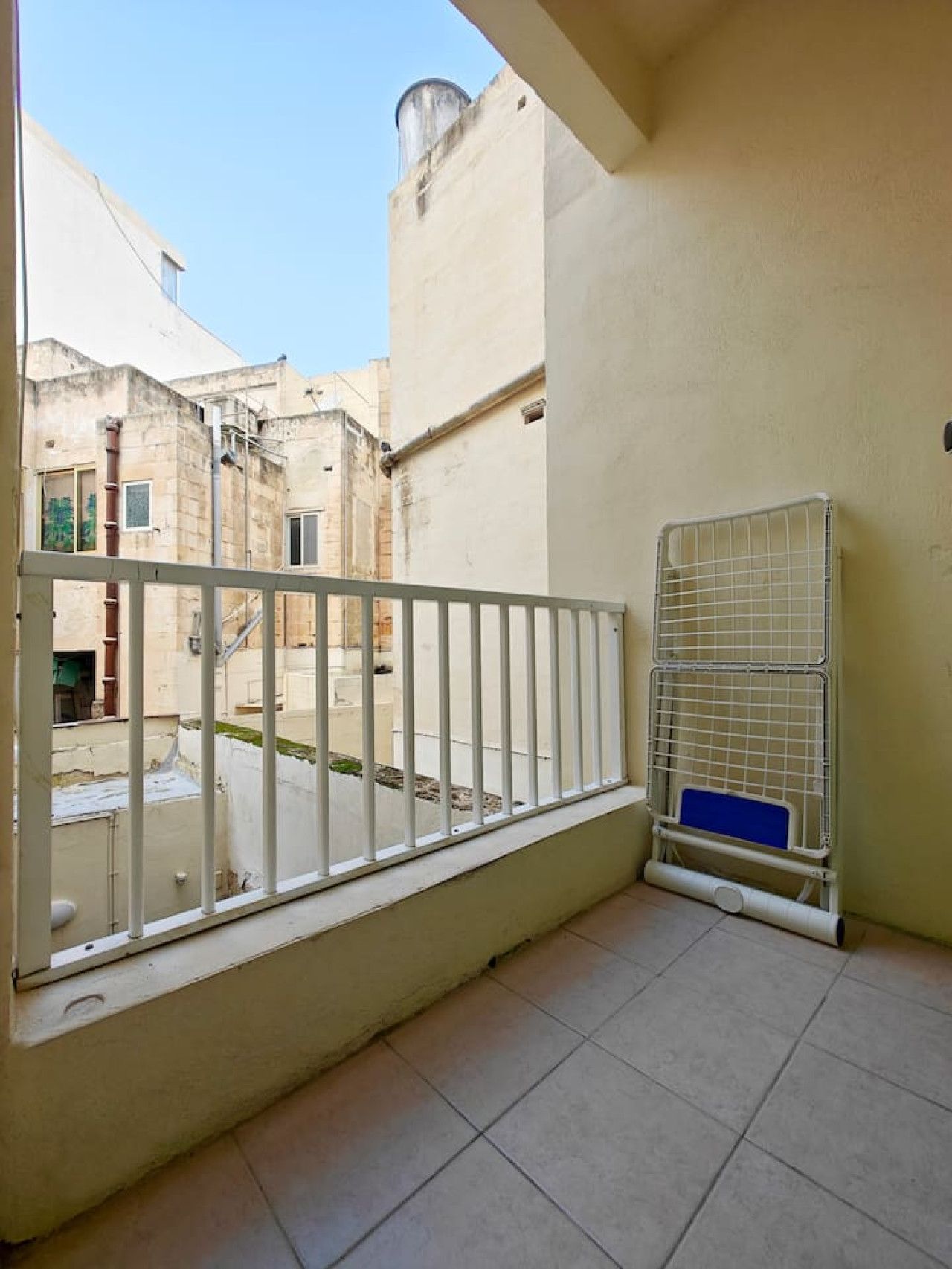 Ein Balkon mit Metallgeländer, ein Wassertank am Nachbarhaus und ein Metallkäfig mit einer blauen Auflage auf dem Boden.