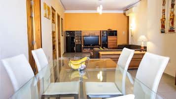 A dining area with a glass table, white chairs, a laptop, and a banana bowl.