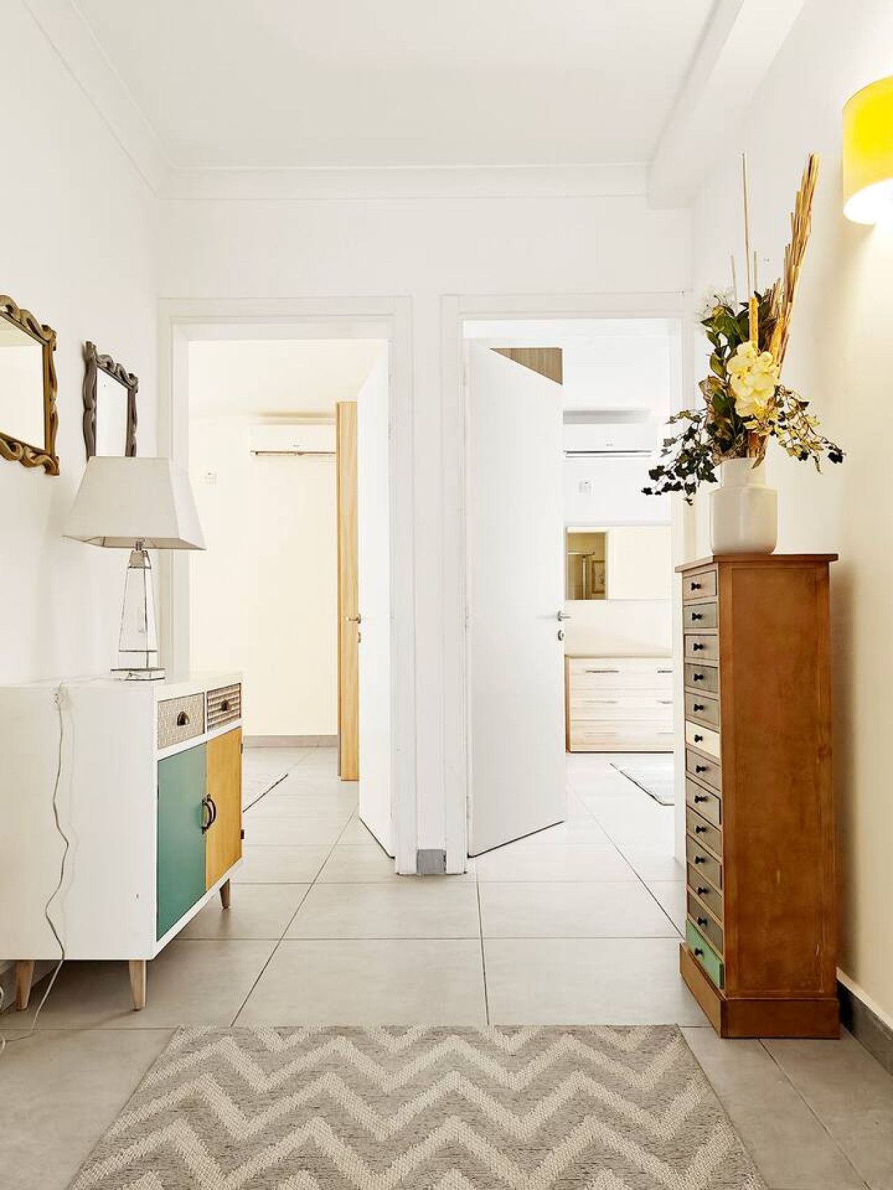 A hallway with a white floor, a herringbone-patterned rug, a white cabinet with a teal door, a wooden cabinet with drawers, and a vase with flowers on a wooden stand.
