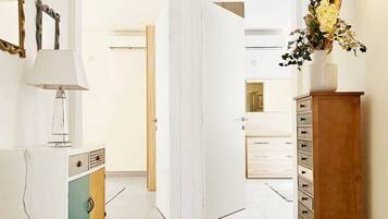 A hallway with a white floor, a herringbone-patterned rug, a white cabinet with a teal door, a wooden cabinet with drawers, and a vase with flowers on a wooden stand.