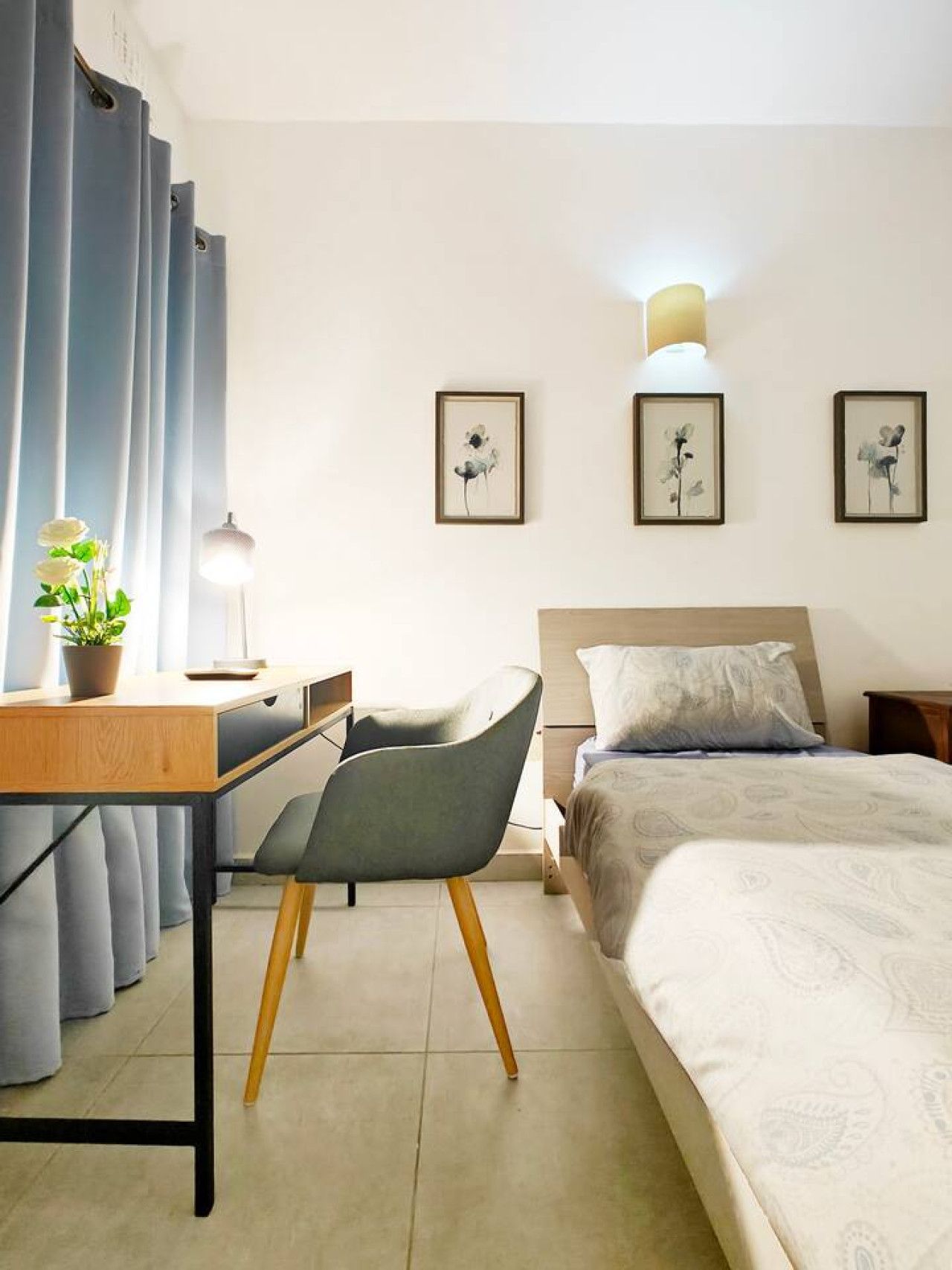 A bedroom with a bed, a desk, a chair, and a bedside table. There are curtains, a lamp, and framed pictures on the wall.