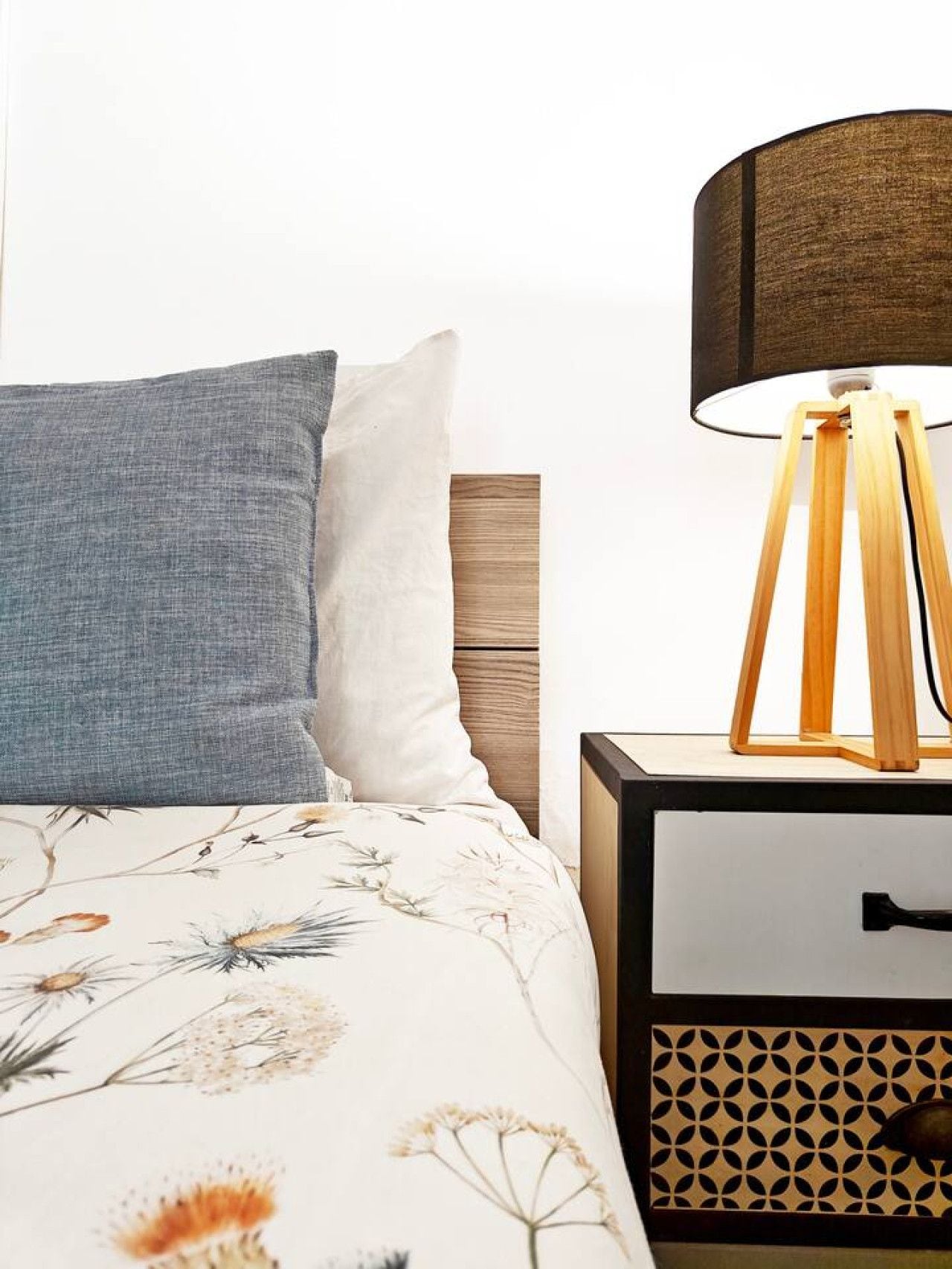 A bed with a floral patterned bedspread, a blue pillow, and a wooden nightstand with a lamp.