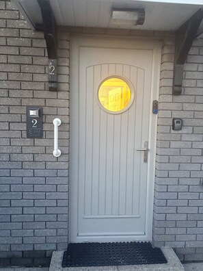 Una porta bianca con una finestra circolare, un campanello e un tastierino su un muro in mattoni.