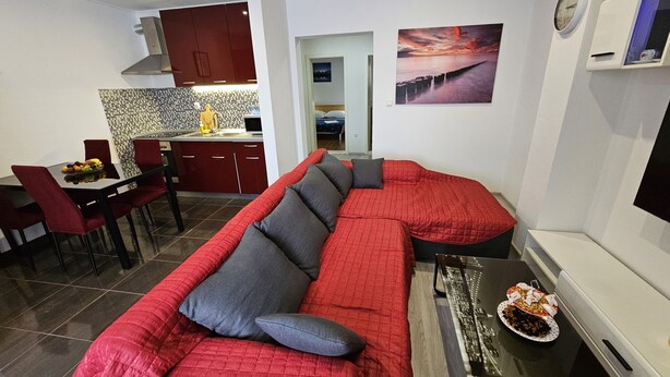 Ein modernes Wohnzimmer mit einem roten Ecksofa, einem Essbereich mit rotem Tisch und Stühlen, einer Küche mit roten Schränken und einem großen Fenster mit Sonnenuntergangsblick.