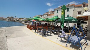 Ein Café am Wasserufer mit Außenbereich, grünen Sonnenschirmen und Gebäuden aus Stein im Hintergrund.