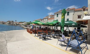 Un caffè sul lungomare con tavolini all'aperto, ombrelloni verdi e edifici in pietra sullo sfondo.