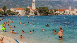 Eine Küstenstadt mit blauem Meer, Menschen beim Schwimmen und Sonnenbaden sowie historischen Gebäuden mit einem markanten Turm.