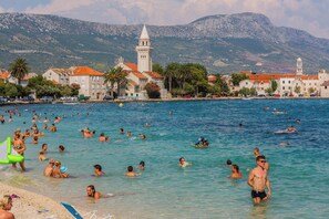Une ville côtière bordée d’une mer d’un bleu éclatant, où l’on peut voir des gens se baigner et bronzer, et où se dressent des bâtiments historiques, dont une tour imposante.