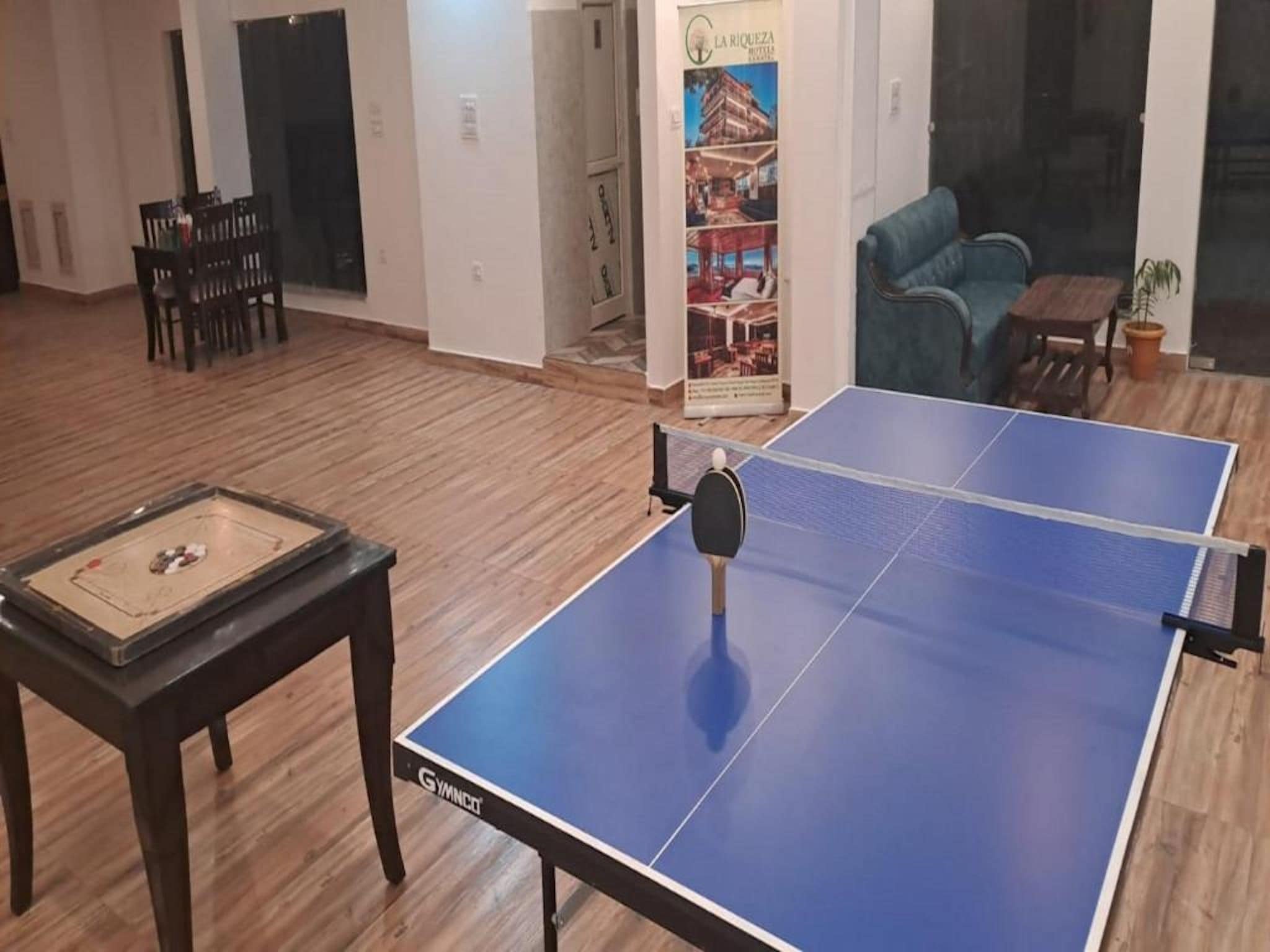 Une table de ping-pong avec une raquette de tennis de table, une petite table avec un jeu de société et une affiche accrochée au mur.