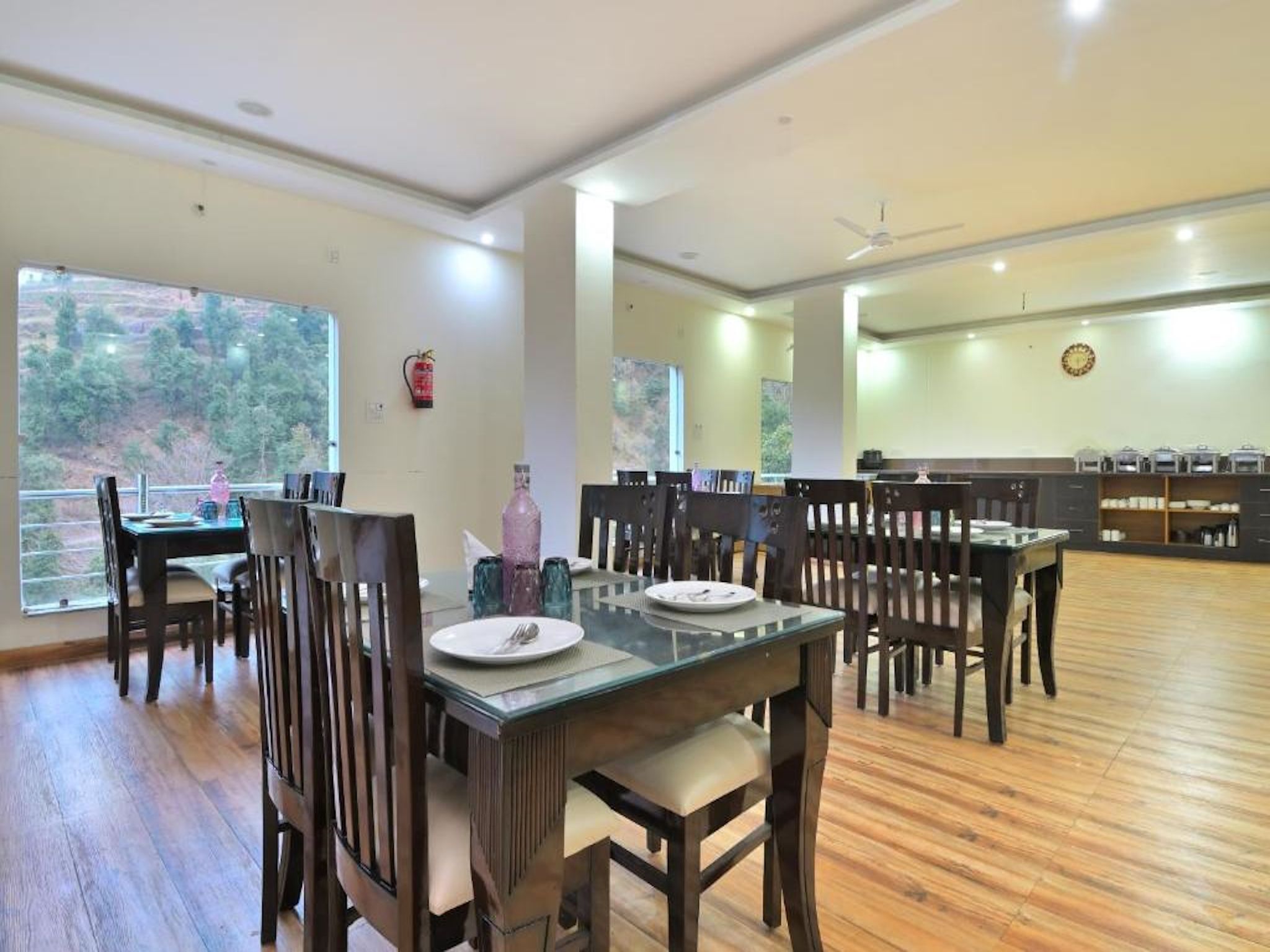 A dining area with wooden tables and chairs, a fire extinguisher on the wall, and a view of the outdoors through large windows.