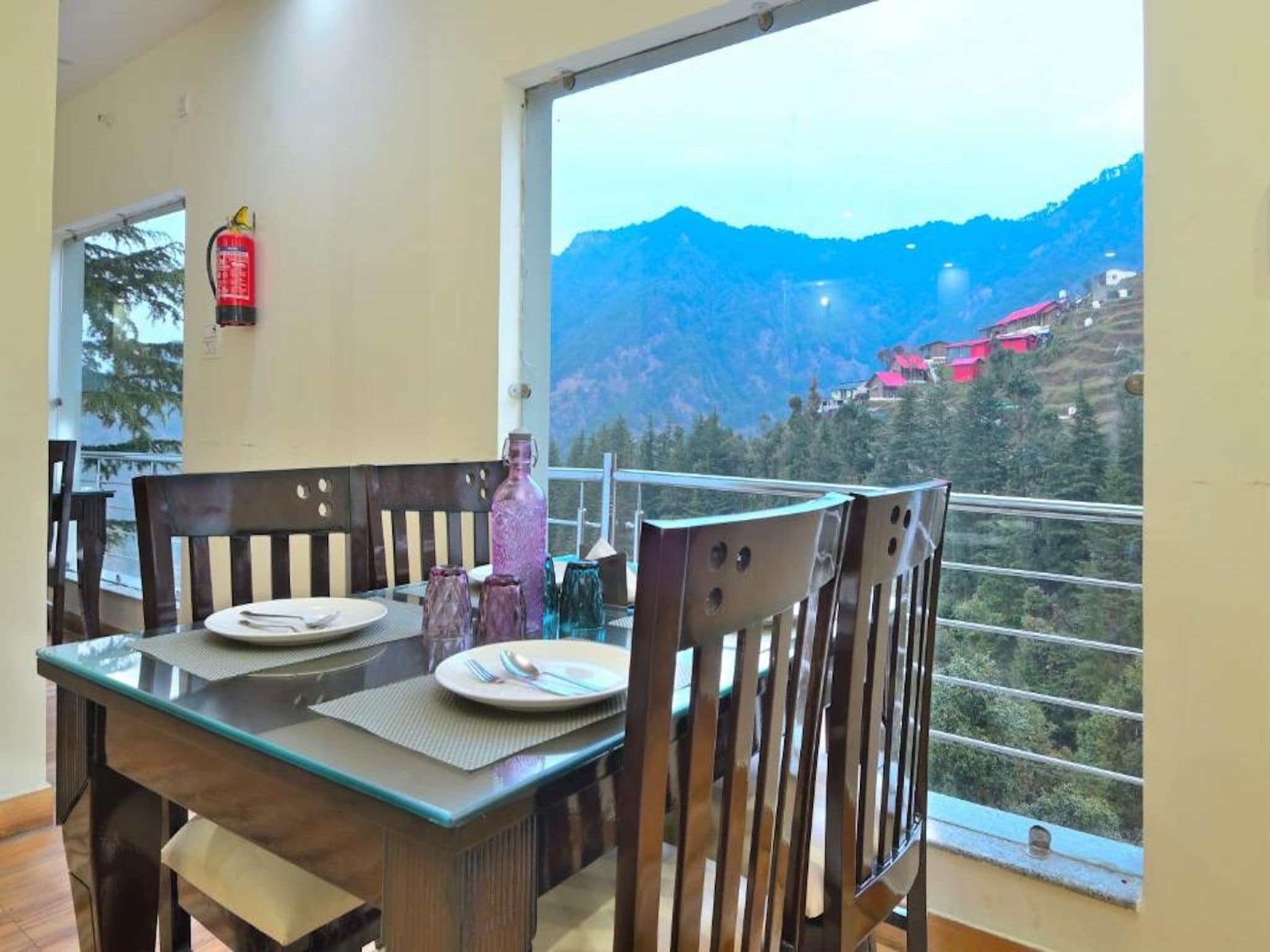 Un comedor con una mesa dispuesta para dos, sillas, un extintor y una ventana con vista a la montaña.