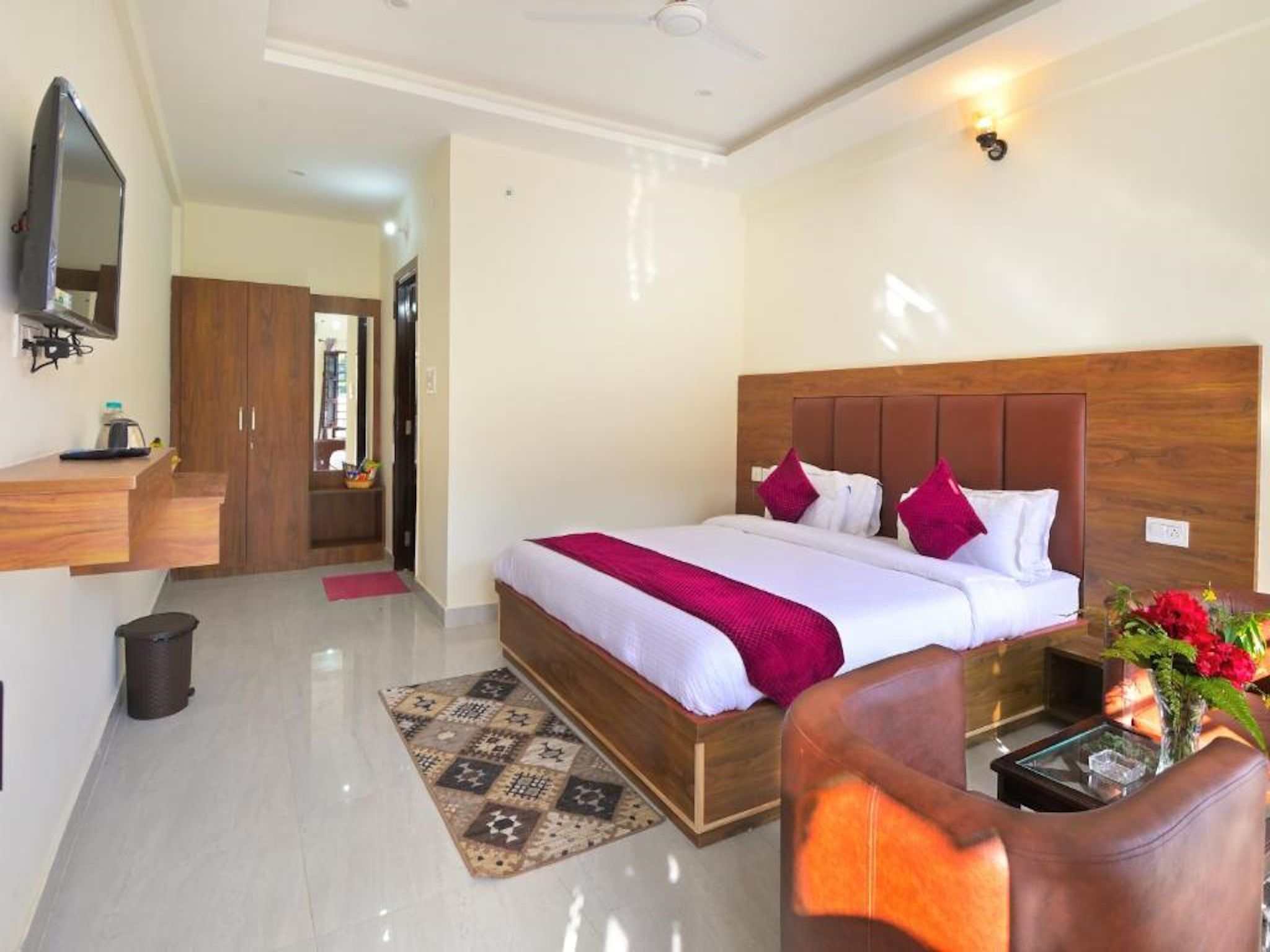 A hotel room with a bed, a TV mounted on the wall, a desk with a chair, and a small table with a vase of flowers.