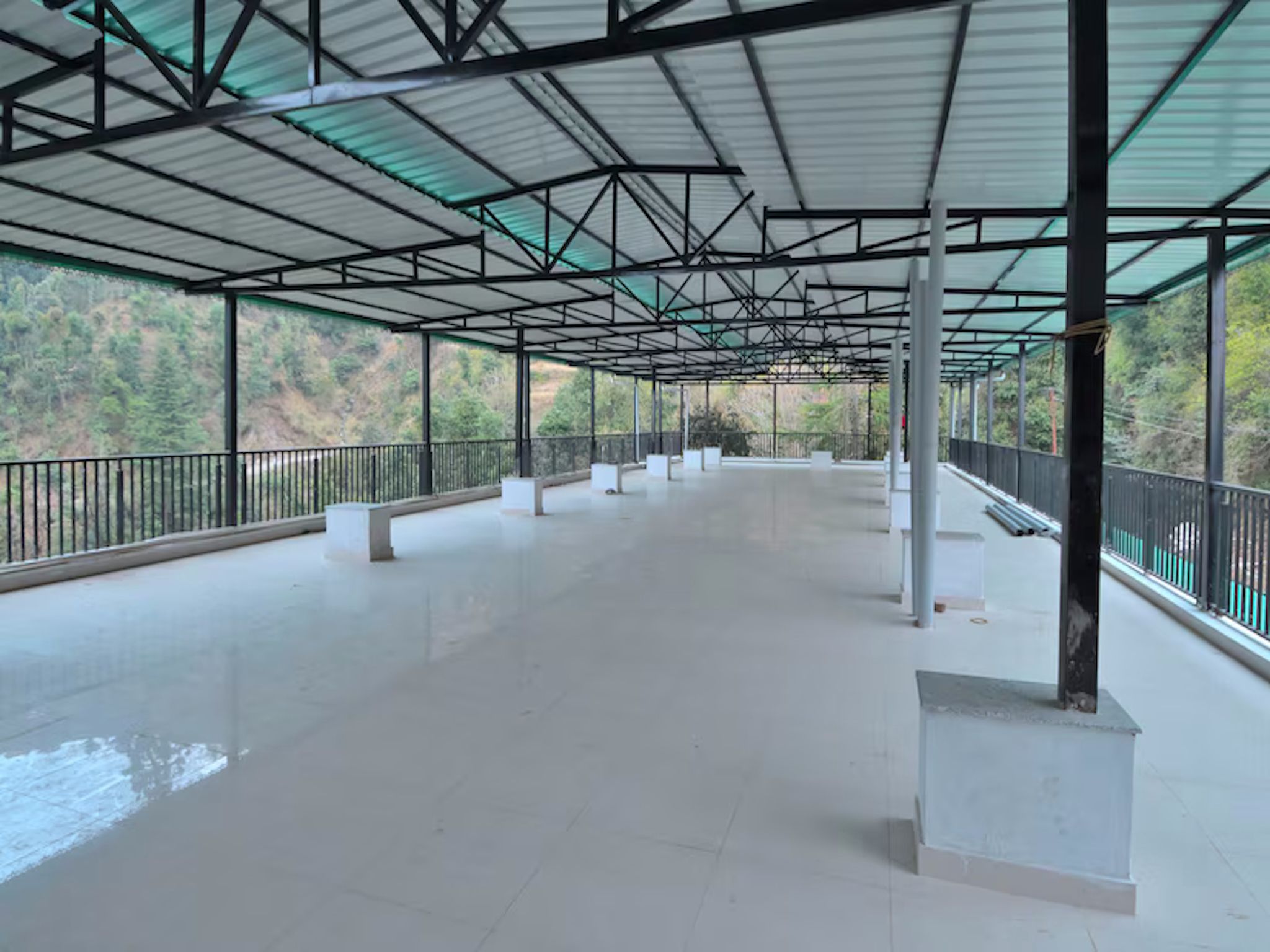 An empty covered walkway with a metal roof and railings, surrounded by greenery.