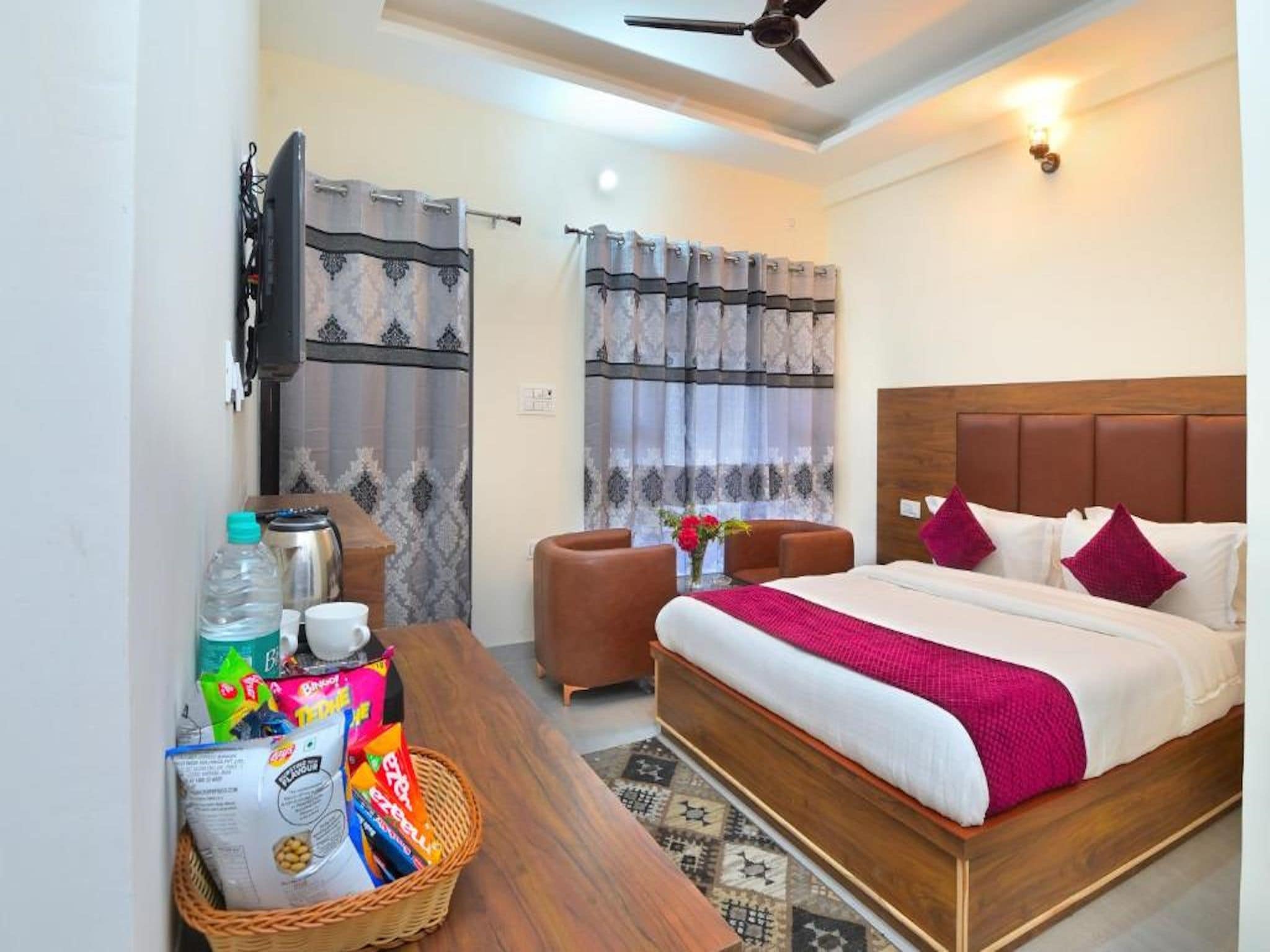 A hotel room with a large bed, a wooden headboard, a desk with a basket of snacks, a coffee maker, and a ceiling fan.