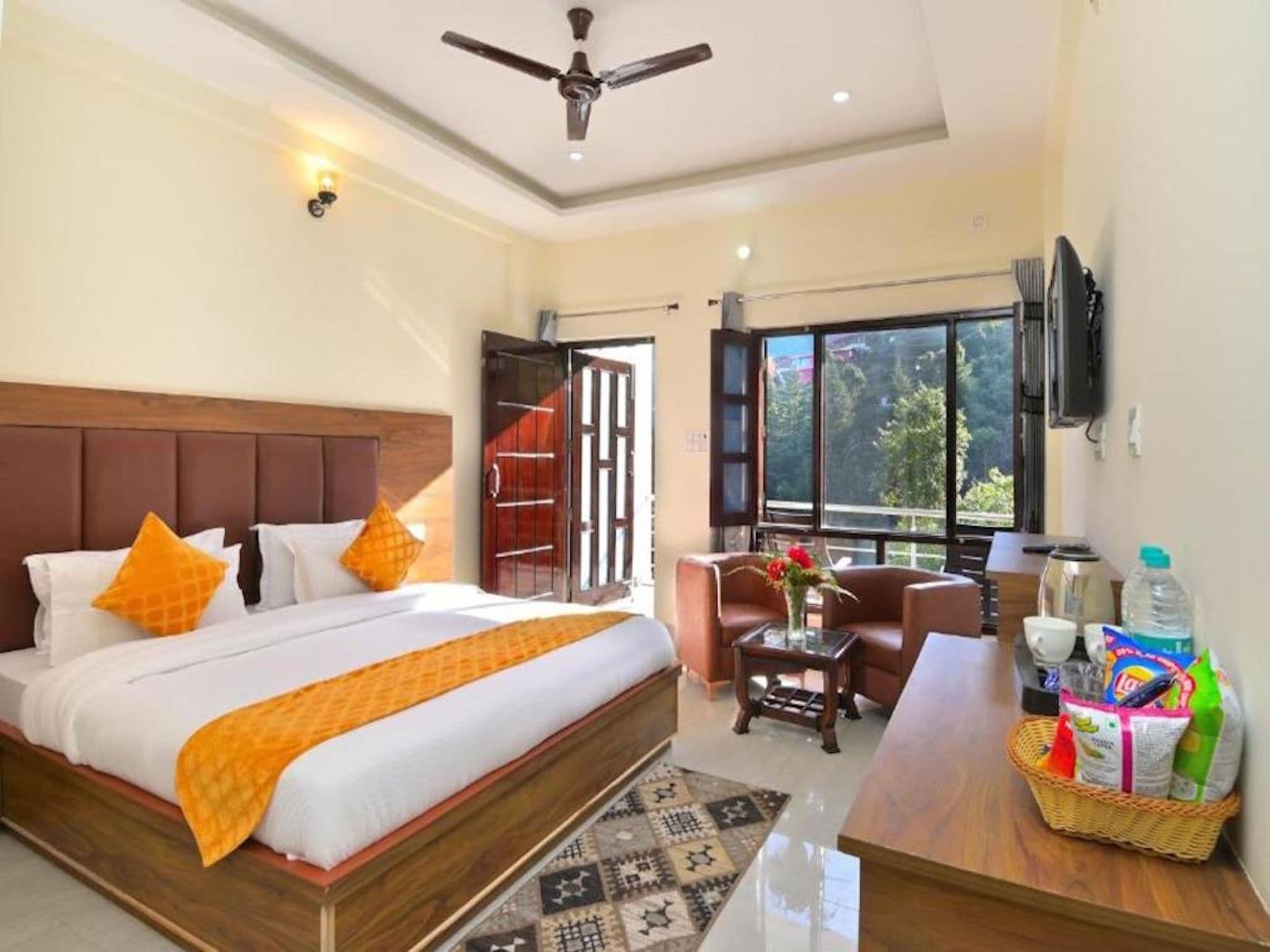 Une chambre d’hôtel avec un grand lit, un ventilateur de plafond, une télévision, une fenêtre avec vue et une petite table sur laquelle se trouve un panier rempli de divers objets.